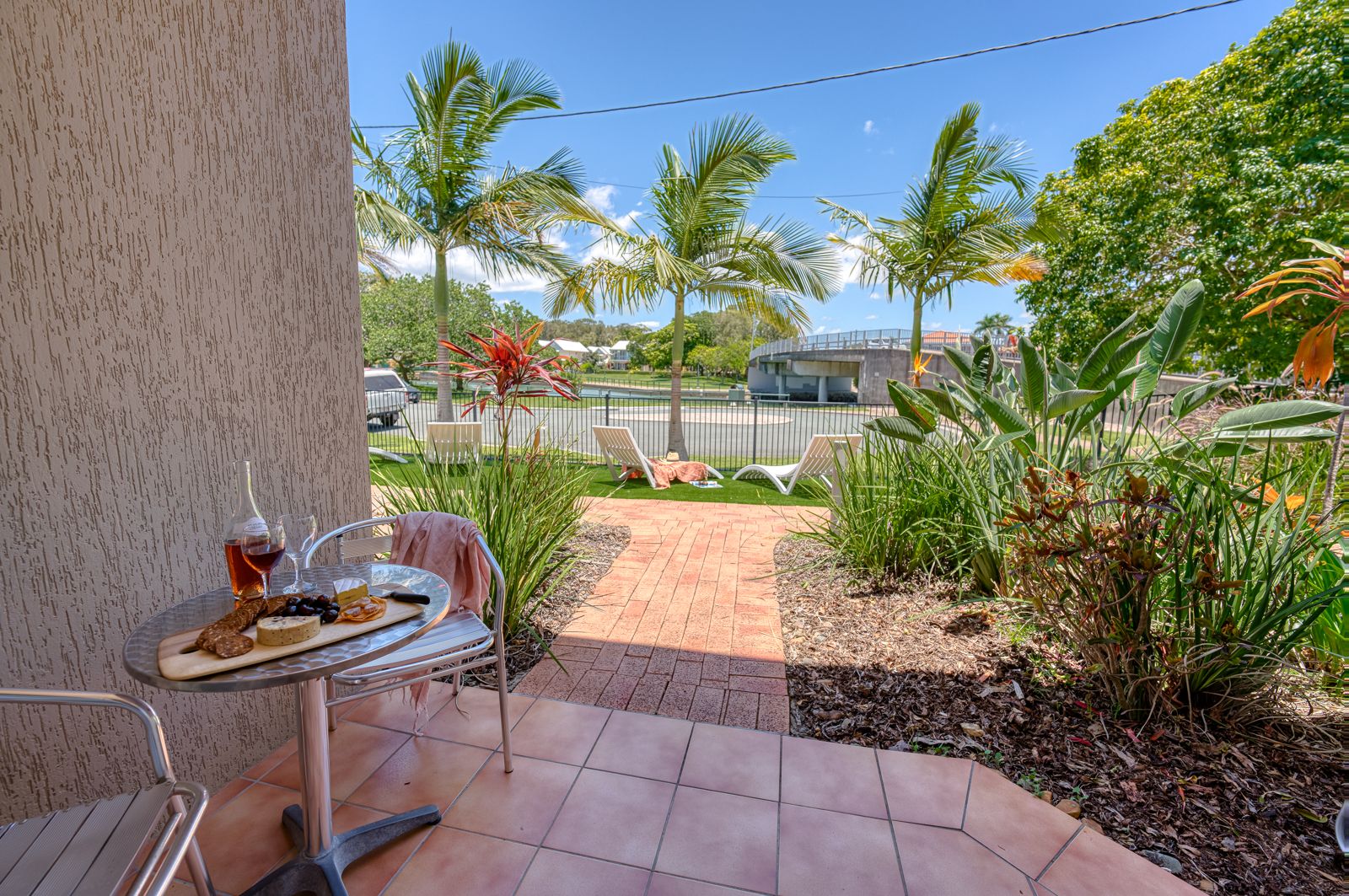 Tiled courtyard with bistro table, opening onto a garden path, lawn and sun loungers, with the canal and bridge beyond